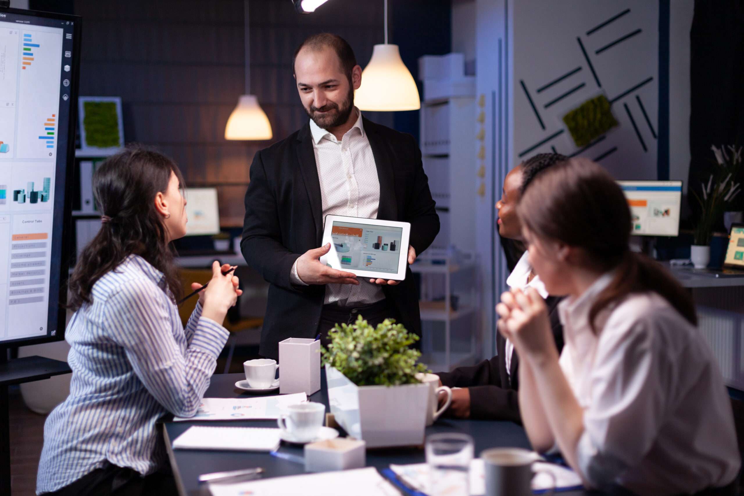 Entrepreneur businessman showing company strategy using tablet for corporate presentation working overtime in meeting office room late at night. Diverse multi-ethnic teamwork brainstorming ideas.